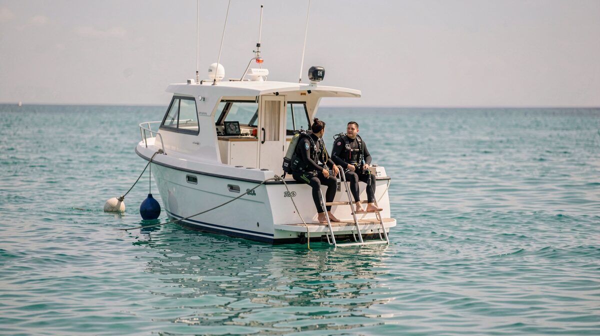 A small white dive boat tied to a mooring buoy on flat turquoise water, stern ladder down, two divers in black wetsuits sitting on the transom, late morning light, shot from water level about ten meters off the bow.